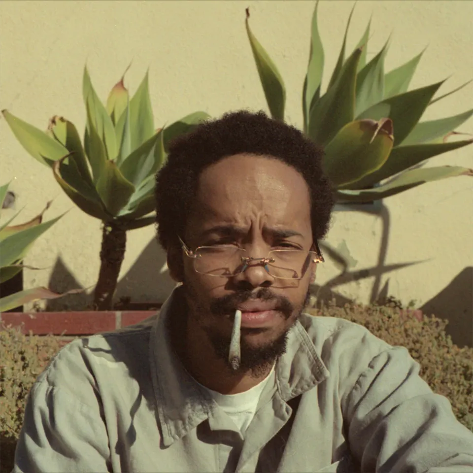 The cover of 'Live, Laugh, Love'. Earl Sweatshirt squinting at the camera with a lit joint hanging precariously from his lips. He's sitting in front of plants outside.